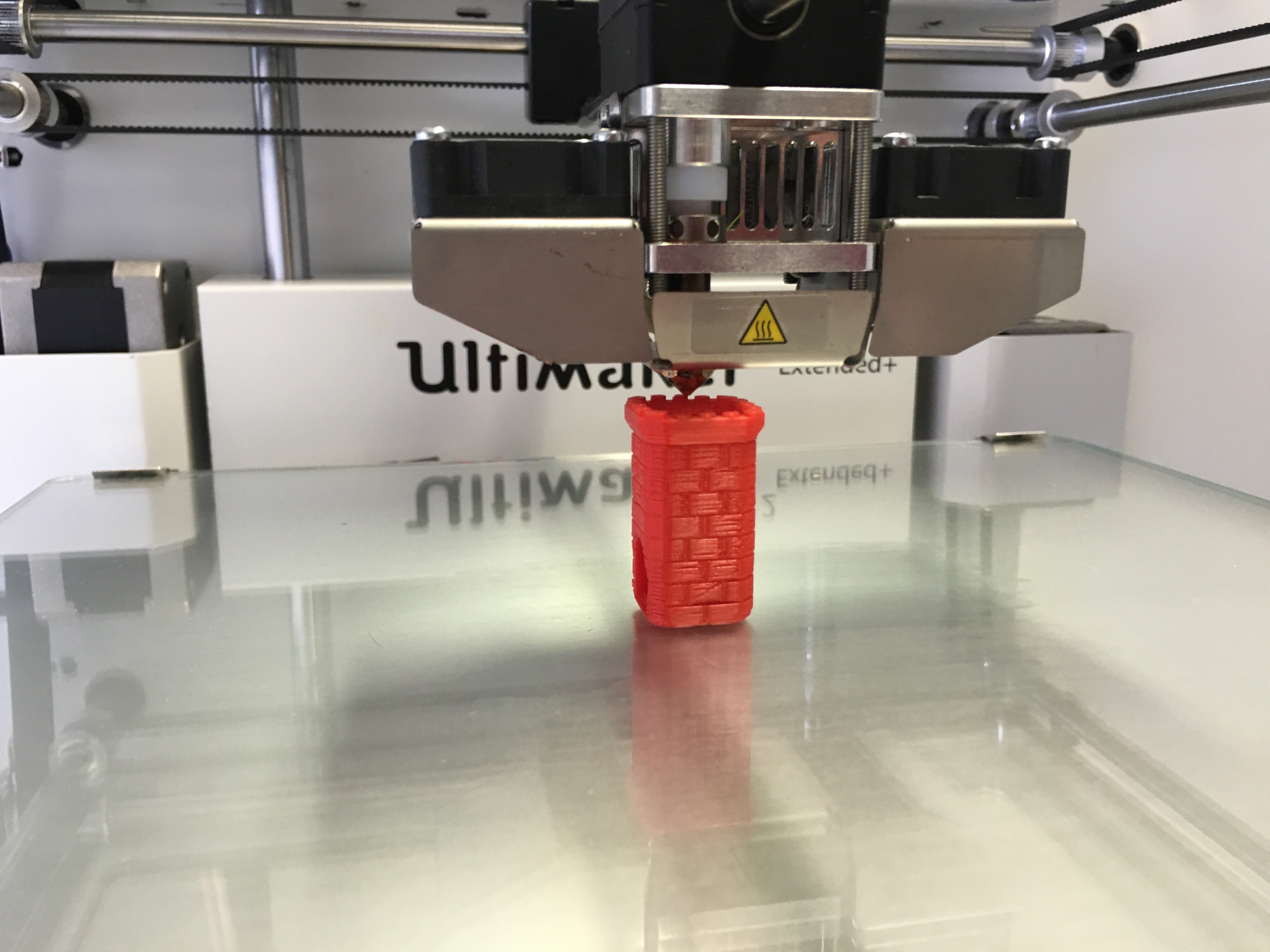 3D printer producing a small red PETG part on a glass print bed